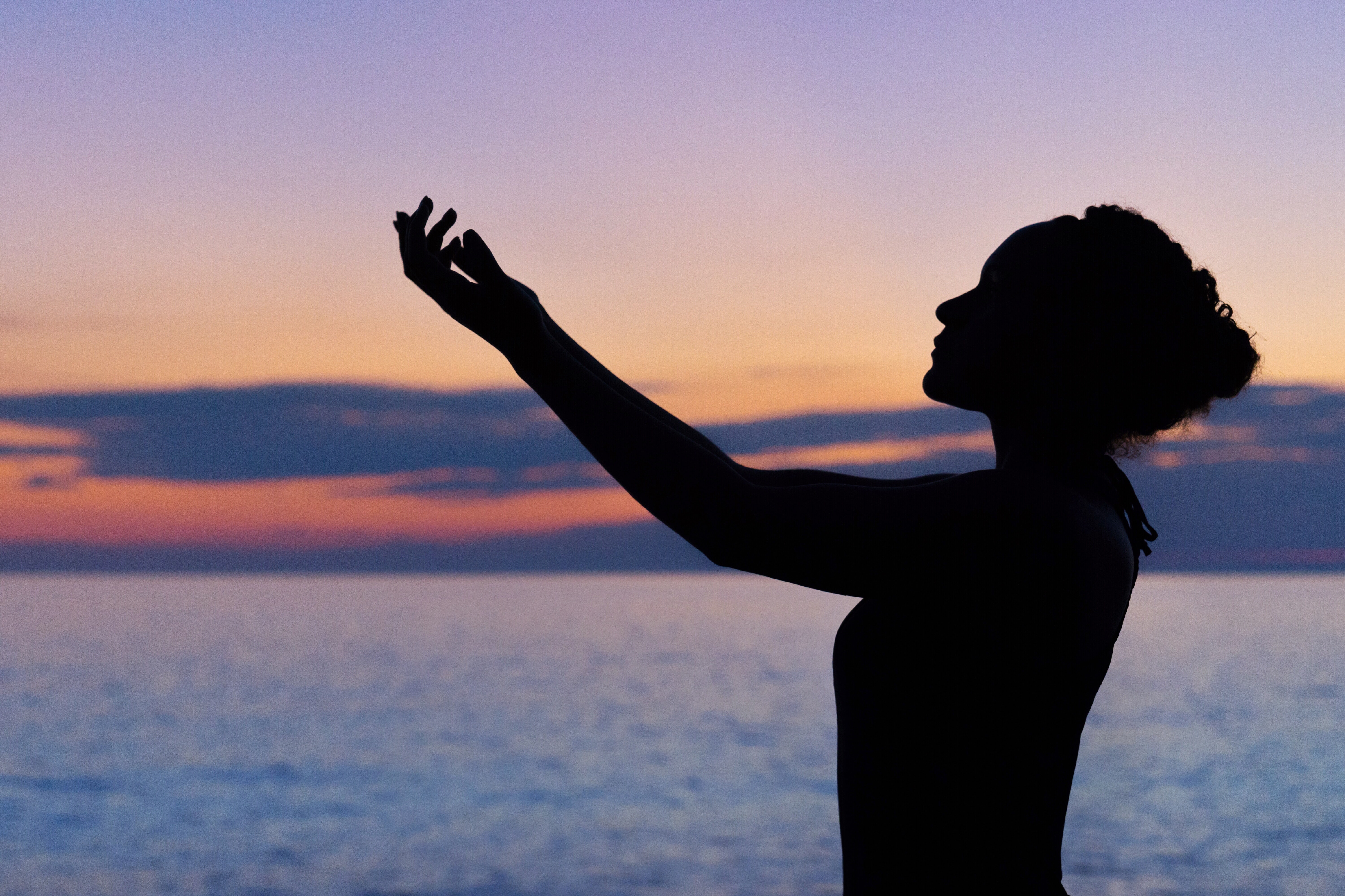 silhouette of a woman with arms raised to the sky, water in the background