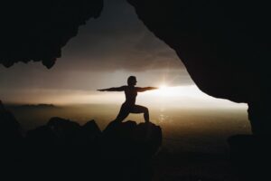 silhouette of a person in warrior two seen from a cave with the setting sun blanketed by clouds