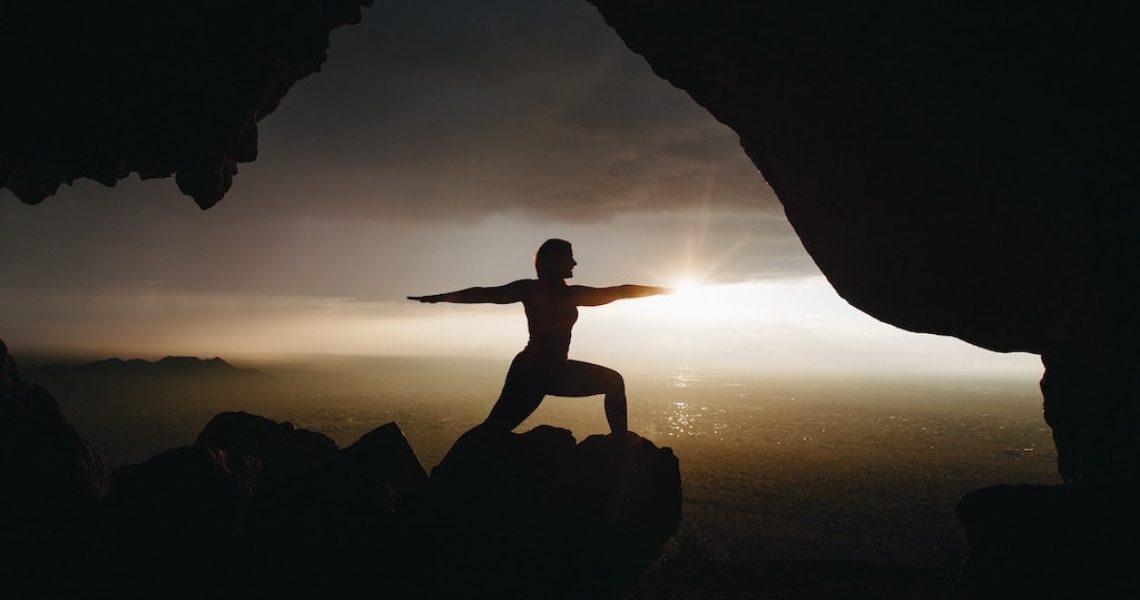 silhouette of a person in warrior two seen from a cave with the setting sun blanketed by clouds