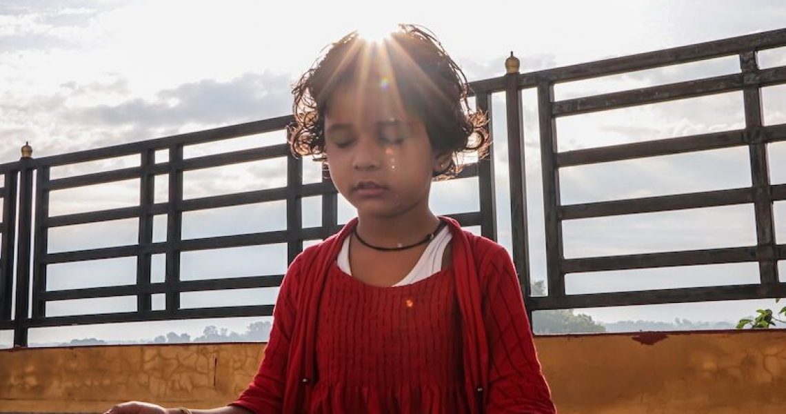 young child sitting in meditation with bright sun shining on their head, she wears a bright red sweater and has a railing in the background