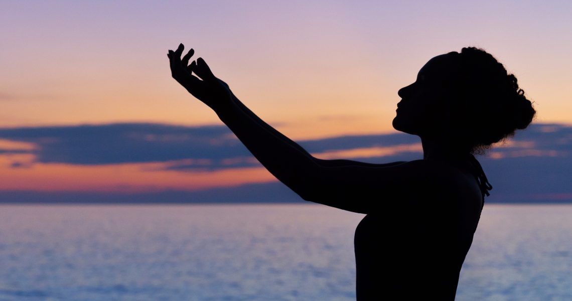 silhouette of a woman with arms raised to the sky, water in the background